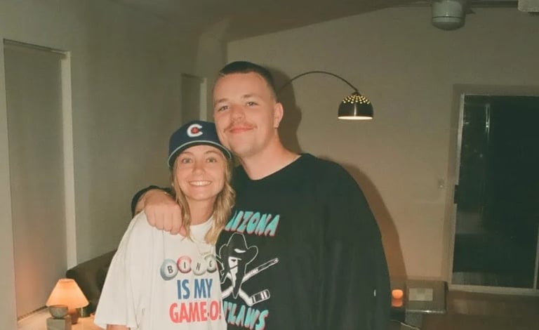 Two people posing together indoors, woman wearing Cubs cap and Coors shirt, man wearing black Arizona shirt, standing with arms around each other